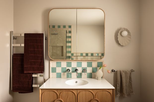 A warm, retro-inspired bathroom vanity vignette featuring a rounded rectangular mirror with a timber-toned frame, a white countertop and integrated basin, and a fluted or carved timber vanity cabinet. The backsplash and reflected shower wall use a checkerboard pattern of cream and muted teal tiles. A chrome wall-mounted tap set, cream wall sconces, towel rails, and dark burgundy and taupe towels complete the softly lit, vintage-styled space.