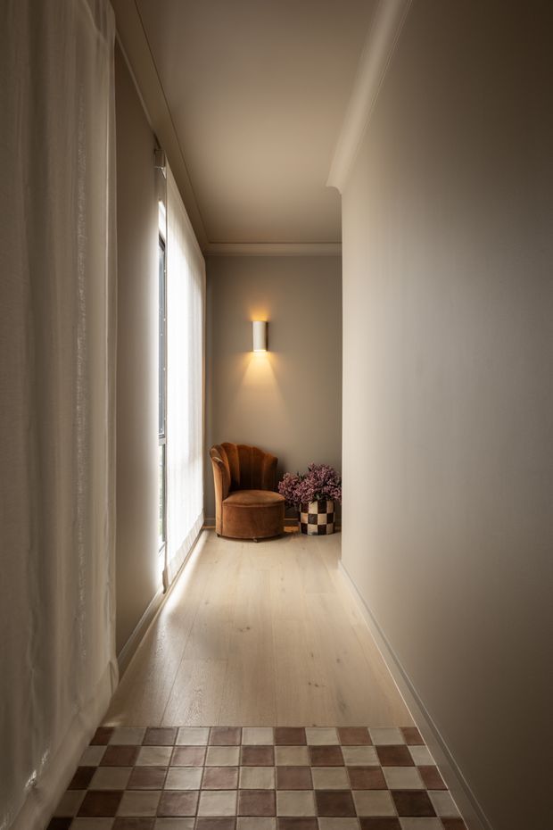 A narrow, warmly lit residential hallway or landing with a long glazed window concealed by sheer curtains on the left, pale timber flooring, smooth warm beige walls, and decorative ceiling cornice. At the far end is a sculptural rust-brown upholstered armchair beside a checkerboard planter with flowers, illuminated by a wall sconce. The foreground has a distinctive brown-and-cream checkerboard tiled threshold.