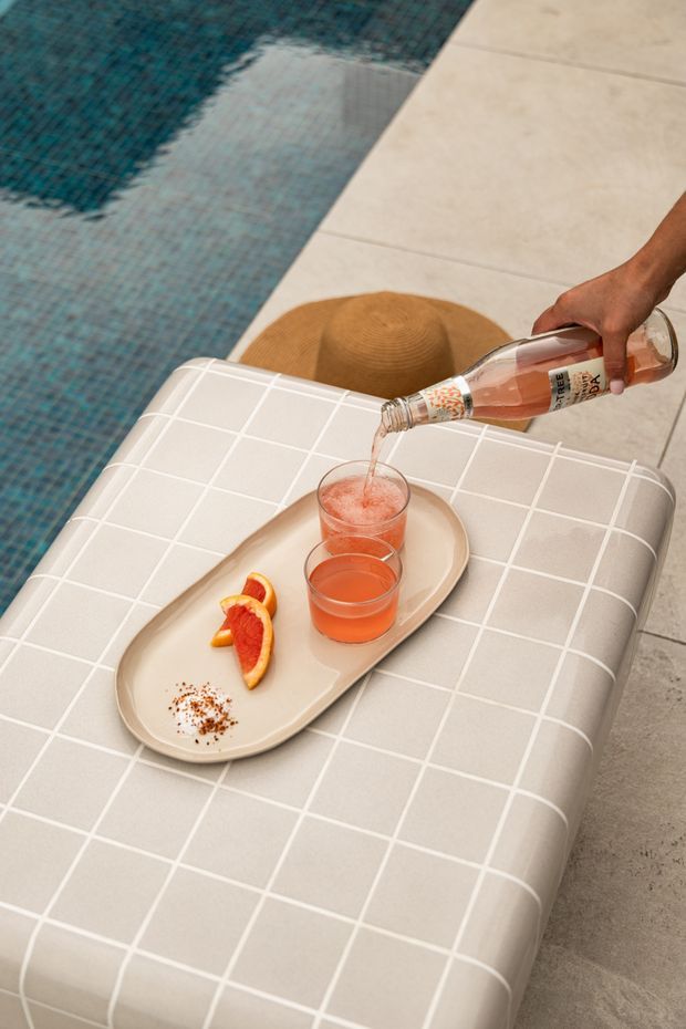 Close-up lifestyle photograph of a poolside setting, showing a pale tiled pool edge, turquoise mosaic swimming pool water, and a cushioned outdoor lounger with a beige grid pattern. A tray holds two pink drinks and sliced grapefruit while a person pours from a bottle. A straw hat rests nearby.
