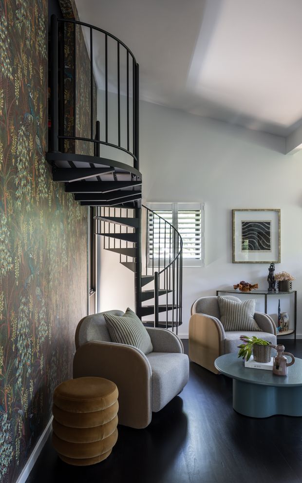 Refined living area with a sculptural black spiral staircase, patterned feature wallpaper, dark timber flooring, and two upholstered lounge chairs arranged around a rounded coffee table. White walls, plantation shutters, curated artwork, and a slim console create a sophisticated, moody interior.