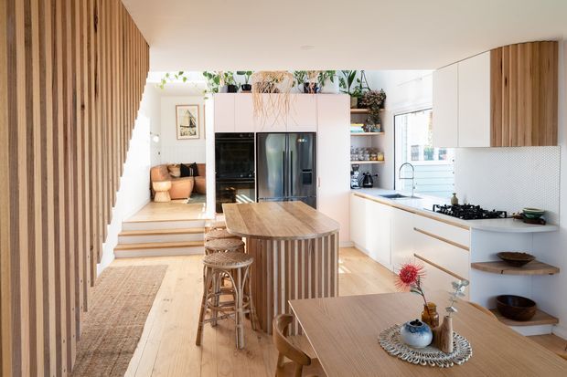 Bright open-plan kitchen and dining area with pale timber flooring, white cabinetry, a central island clad in vertical timber battens, timber wall screening, integrated appliances, open shelving, and a small raised sitting area beyond. Natural light and indoor plants create a warm, airy atmosphere.