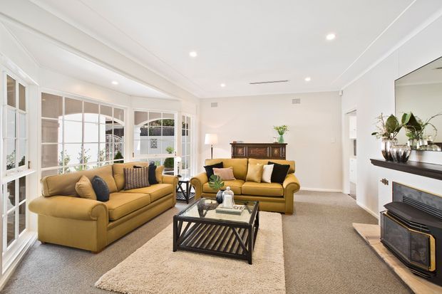 Bright, traditionally styled living room with two mustard-gold upholstered sofas, a glass-topped black coffee table, beige carpet and rug, white walls and ceiling, extensive white-framed glazed doors and windows, and a fireplace with dark trim and built-in shelving. A timber sideboard and decorative furnishings add warmth to the room.