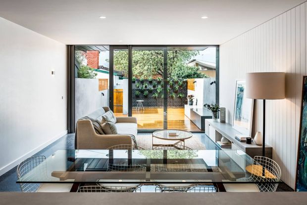 A bright, contemporary living room with a glass dining table and wire-frame dining chairs in the foreground, a neutral sofa and round coffee table, and a long built-in media console along a vertically panelled wall. Full-height glazing opens toward a small timber-decked courtyard or garden, bringing in abundant natural light.