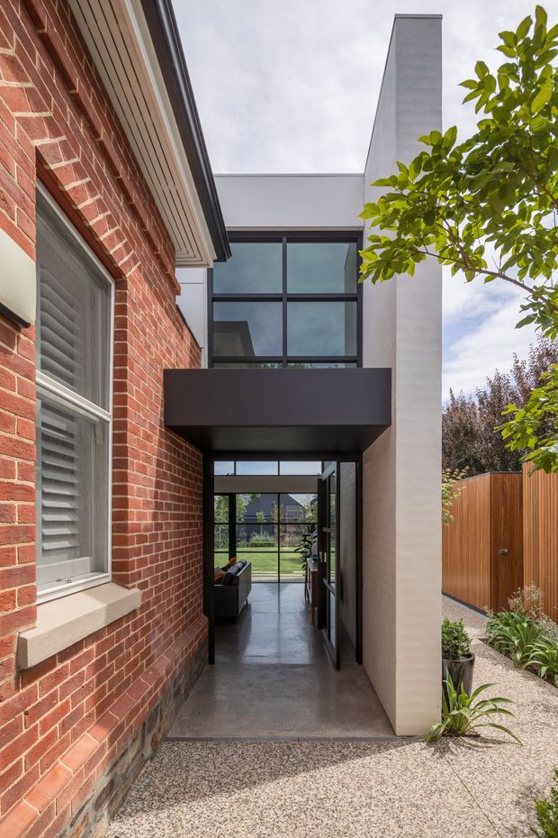 Photograph of a contemporary residential entry passage viewed from outside, combining an existing red brick facade with a sharply contrasting modern addition. The addition features a tall white rendered wall, charcoal metal canopy, expansive black-framed glazing, and a glazed door opening into a dark polished interior. A textured aggregate-paved path, timber screening fence, and planted garden edge frame the entrance.