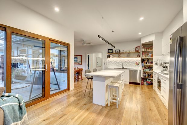Bright, open-plan kitchen with a central island and bar stools, white cabinetry, a patterned backsplash, stainless-steel appliances, and warm timber flooring. Large timber-framed glazed doors open toward an outdoor area, while a dining space is visible beyond the kitchen.