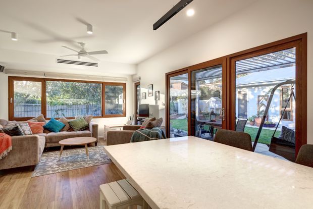 Bright open-plan living and dining area viewed from a kitchen island. The room features warm timber flooring, a large light stone island benchtop, white walls and ceiling, timber-framed glazing, comfortable upholstered seating, and extensive glass doors opening to a landscaped courtyard. Contemporary linear track lighting and a ceiling fan complement the relaxed, light-filled interior.