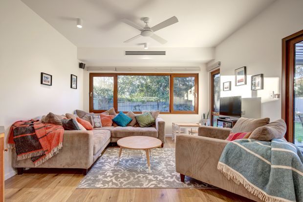 A bright, comfortably furnished living room with large timber-framed windows and glazed doors opening toward greenery outside. The room features a large L-shaped sectional sofa, matching armchairs, patterned area rug, round timber coffee table, television and low media unit, framed wall art, ceiling fan and recessed lighting. Warm timber flooring and layered neutral textiles create an inviting, relaxed atmosphere.