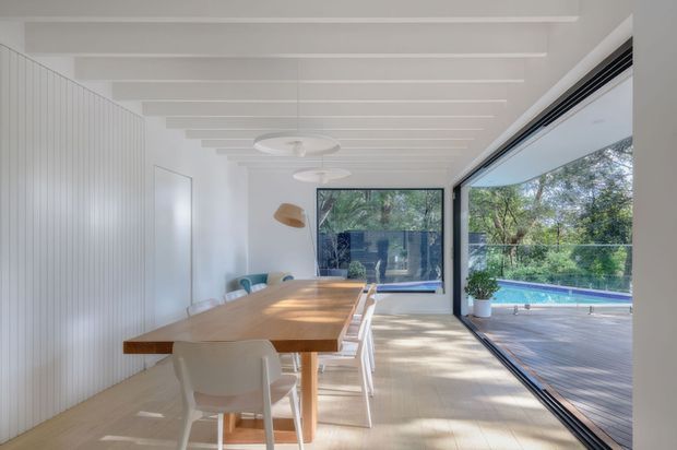 Bright contemporary dining area with a long light timber dining table and white chairs, enclosed by full-height glazing opening to a timber deck and swimming pool. The room features white vertical wall panelling, exposed white ceiling battens, pale flooring, integrated linear glazing, and sculptural pendant lights.