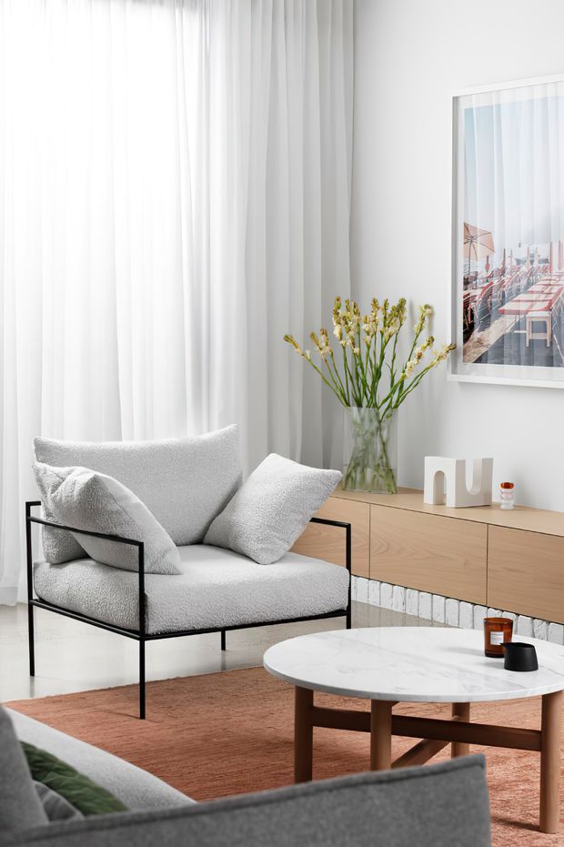 Bright, refined living room with a white boucle armchair, grey sofa in the foreground, light timber sideboard, round marble coffee table, sheer floor-to-ceiling curtains, framed artwork, and a low white-painted brick feature wall. The palette is predominantly white, light grey, pale timber, and muted terracotta.