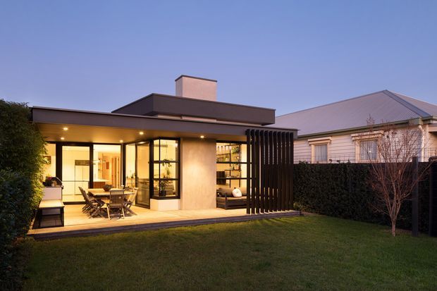 Twilight exterior view of a contemporary single-storey backyard extension or pavilion. The building has a flat roof with a raised rectangular chimney or roof element, warm interior lighting, extensive glazed sliding doors, a timber deck with outdoor dining furniture, and a dark vertical slatted screen. It sits beside a pitched-roof weatherboard residence, with a lawn and hedging in the foreground.