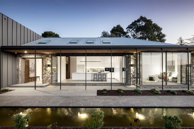 A low, contemporary single-storey residence viewed from across a narrow landscaped pond. The home has extensive floor-to-ceiling glazing, a broad metal-clad roof with several skylights, dark slender window frames, stone-clad structural piers, and a covered outdoor walkway or terrace. A bright minimalist kitchen and living area are visible through the glass. The foreground includes a paved path and reflective water feature with planting.