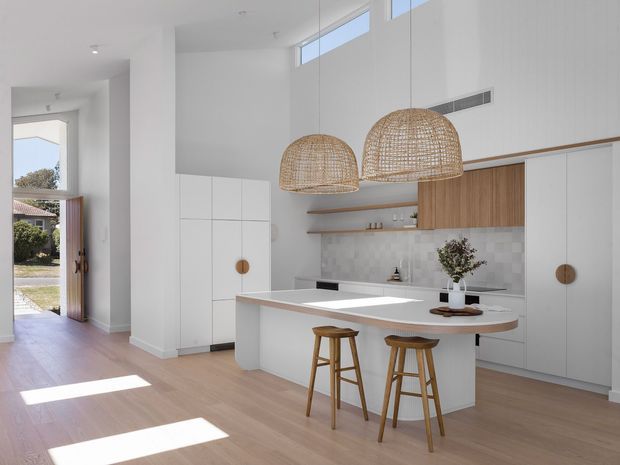 Bright open-plan kitchen with a large white island, timber stools, white integrated cabinetry, warm timber feature joinery, pale tiled splashback, and woven pendant lights. The kitchen opens toward an entry hall and exterior doorway, with high-level glazing bringing in abundant natural light.