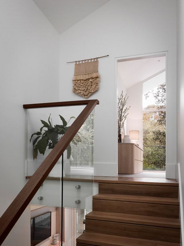 A bright, minimalist stair landing with a dark timber handrail and warm timber stair treads. Clear glass balustrades and white walls create an airy, open feel, while a woven wall hanging and indoor plant add natural texture. An open doorway leads to a softly furnished room with large glazing and views of trees.