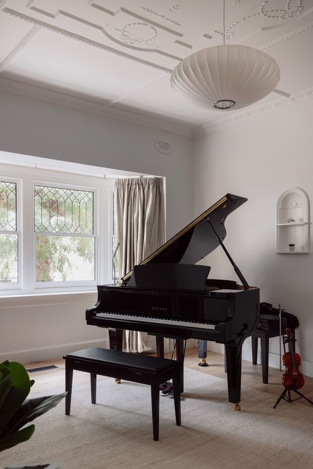 Elegant music room featuring a glossy black grand piano and matching bench, with a violin nearby. The bright room has large leaded-glass windows, layered curtains, ornate decorative ceiling mouldings, a sculptural pendant light, pale walls, and a neutral carpet. A small arched wall niche adds further traditional detailing.