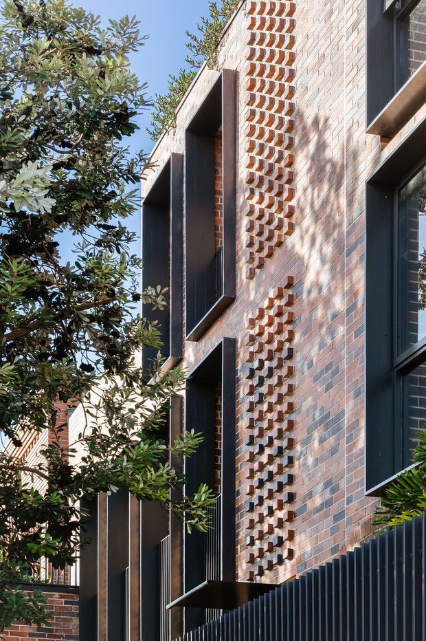 Vertical architectural photograph of a contemporary multi-storey brick facade. The building features patterned projecting brickwork forming a geometric screen, deep dark-framed window and balcony openings, and dark metal privacy fencing. Mature greenery partially frames the facade against a clear blue sky.