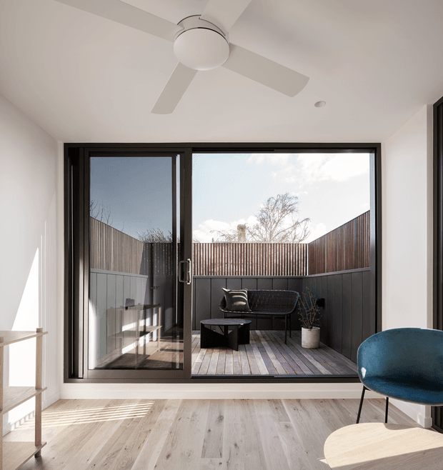 Bright contemporary interior opening onto a compact private courtyard terrace through large black-framed sliding glass doors. The room has pale timber flooring, white walls and ceiling, a minimalist white ceiling fan, and a blue accent chair. Outside, the timber-decked courtyard is enclosed by dark vertical and horizontal privacy fencing and furnished with a black outdoor sofa, coffee table and planter.