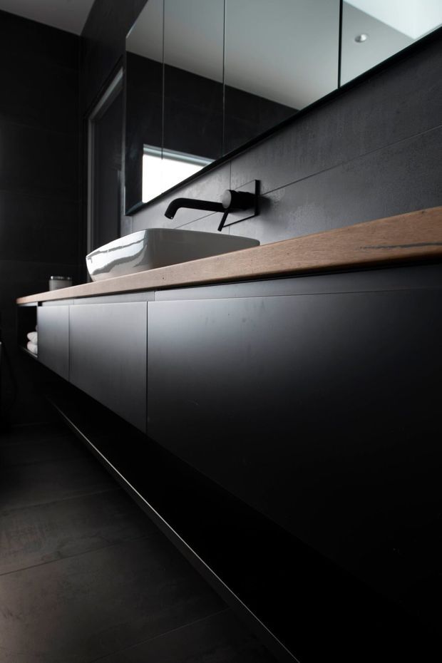 A moody, contemporary bathroom vanity featuring a long floating black cabinet with minimalist flat-panel fronts, a thick warm timber benchtop, a white vessel basin, and a matte black wall-mounted tap. Dark tiled walls and floor, large mirror panels, and concealed or indirect lighting create a dramatic, refined atmosphere.