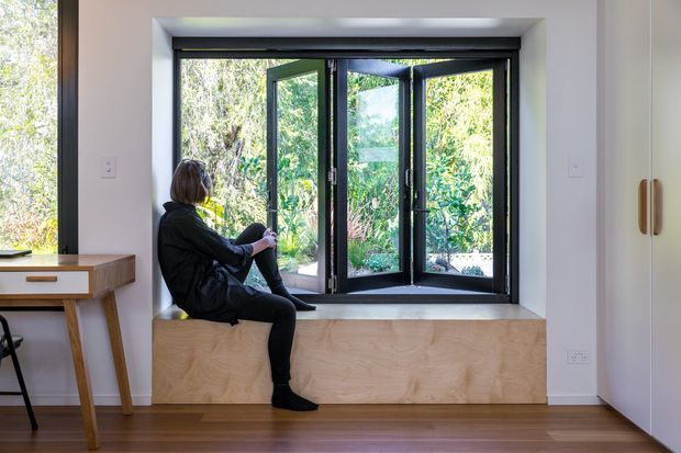 A bright, minimalist interior centered on a deep timber window seat beneath large black-framed bi-fold windows. A person sits on the built-in seat, with leafy vegetation visible outside. The room has white walls, warm timber flooring, light timber joinery, and a compact timber console at the left.