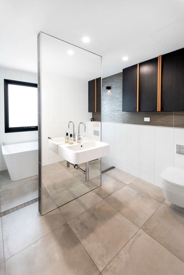 Bright contemporary bathroom featuring a freestanding white bathtub beside a black-framed window, a wall-mounted white basin with exposed plumbing, a frameless mirrored shower partition, wall-mounted toilet, large beige stone-look floor tiles, and dark timber-look cabinetry with warm timber accents.