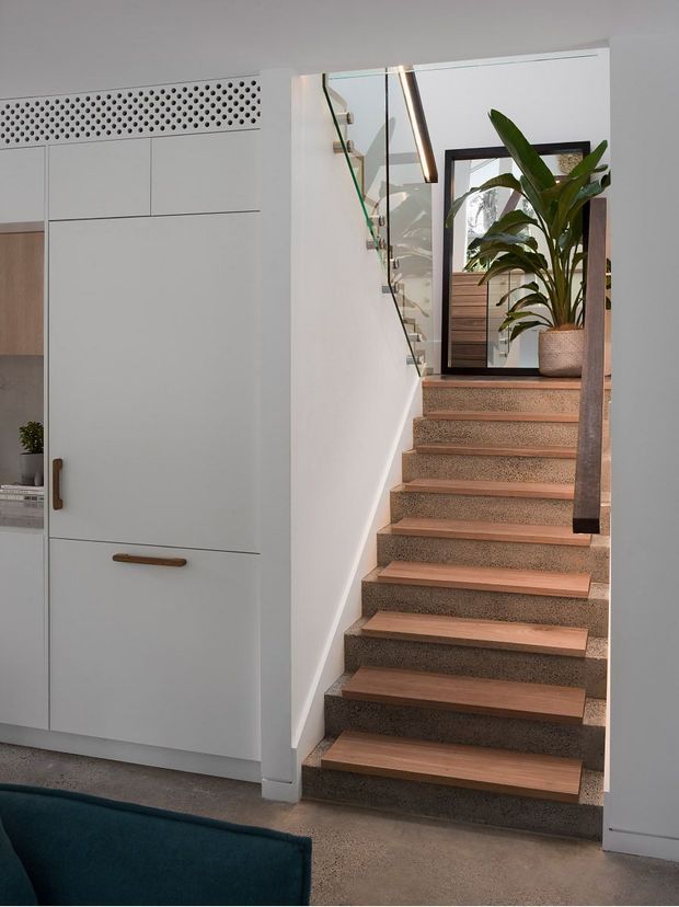 Interior view of a compact contemporary stairway beside full-height white built-in cabinetry. The staircase has exposed timber tread inserts over textured grey stone-like risers, a dark timber handrail/post, and a glass balustrade. A large potted plant and framed mirror sit on the upper landing, while a perforated ventilation panel is integrated above the cabinetry. The palette is neutral with warm timber accents and greenery.