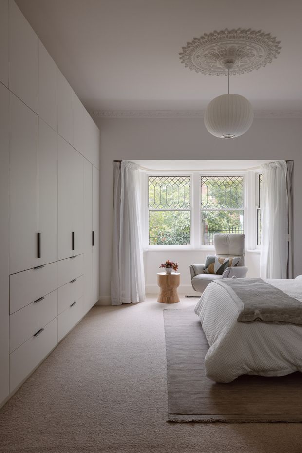 A calm, softly lit bedroom with a large built-in wall of pale cabinetry, a carpeted floor, and a neatly made bed in neutral bedding. A bay window with leaded-glass detailing is framed by sheer white curtains, with a sculptural armchair and small wooden side table beneath an ornate ceiling medallion and pendant light. The room has a refined, restrained traditional character with contemporary built-in storage.