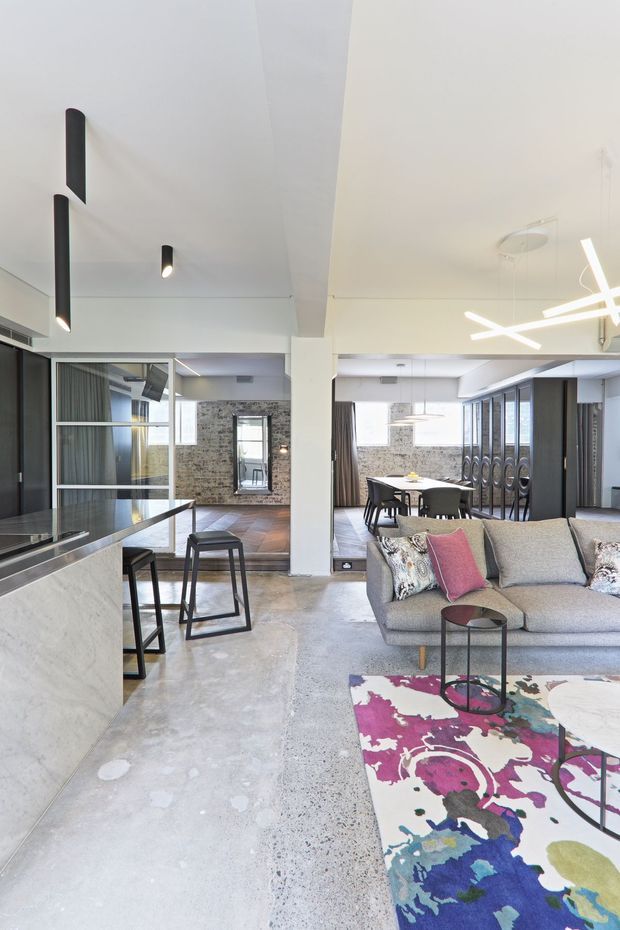 Open-plan contemporary interior combining a kitchen, living area and dining space. The room has polished light concrete flooring, a pale stone kitchen island, dark framed glazing and partitions, a textured exposed brick feature wall, a neutral sectional sofa, and a vivid abstract area rug. Sculptural linear pendant lighting and recessed ceiling lights add a modern finish.