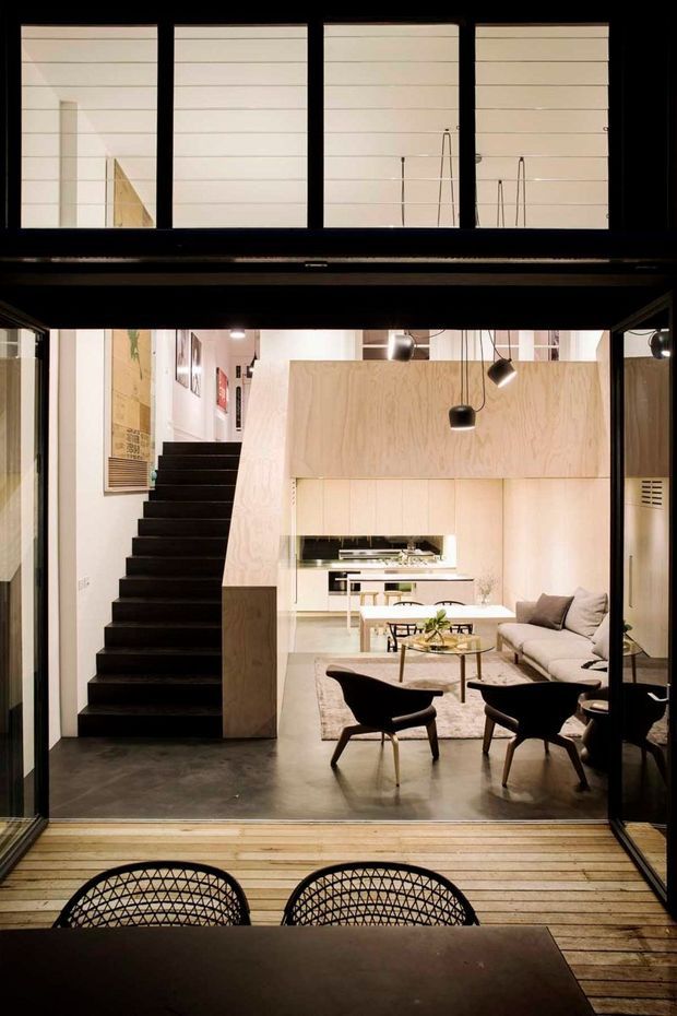 A contemporary open-plan interior viewed through large black-framed glazing from a timber deck. The space features a dark stairway, pale plywood wall panelling and cabinetry, a compact kitchen, a living area with a light sectional sofa, sculptural lounge chairs, pendant and track lighting, and polished grey concrete flooring. Horizontal screened glazing is visible above the room, creating a strong layered architectural composition.
