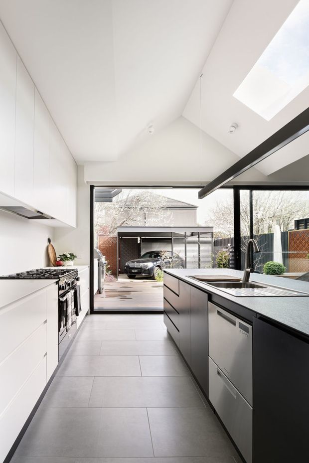 A bright, contemporary galley kitchen with white full-height cabinetry on one side and a dark island with an integrated sink and stainless-steel appliances on the other. Large glazed doors open to a timber deck and carport area, while roof skylights and a high sloping ceiling bring in abundant natural light. The palette is predominantly white, grey, black, and stainless steel.