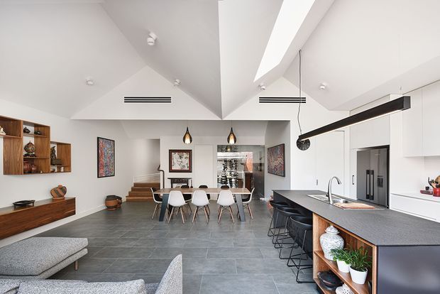 Open-plan contemporary kitchen, dining and living area with a dramatic vaulted ceiling and central raised ceiling beam. The kitchen features white full-height cabinetry, a dark stone-look island benchtop, integrated appliances and timber open shelving. A timber dining table with white chairs sits beside a glass-fronted display cabinet, while grey tiled flooring and neutral upholstered seating create a clean, refined interior. Linear pendant lighting, recessed spotlights and dark accents add contrast.