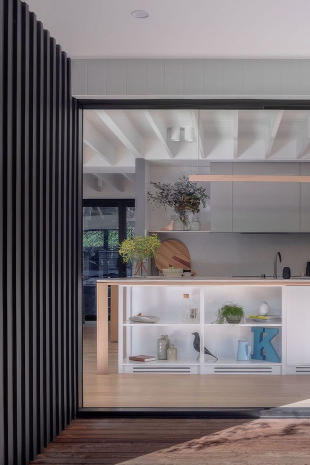 A contemporary kitchen viewed through a wide black-framed opening, with a light timber island featuring open white shelving, pale cabinetry, a grey splashback, timber flooring, and exposed white ceiling beams. A dark vertical slatted screen frames the left side, while plants, decorative objects, and a timber deck add warmth and texture.
