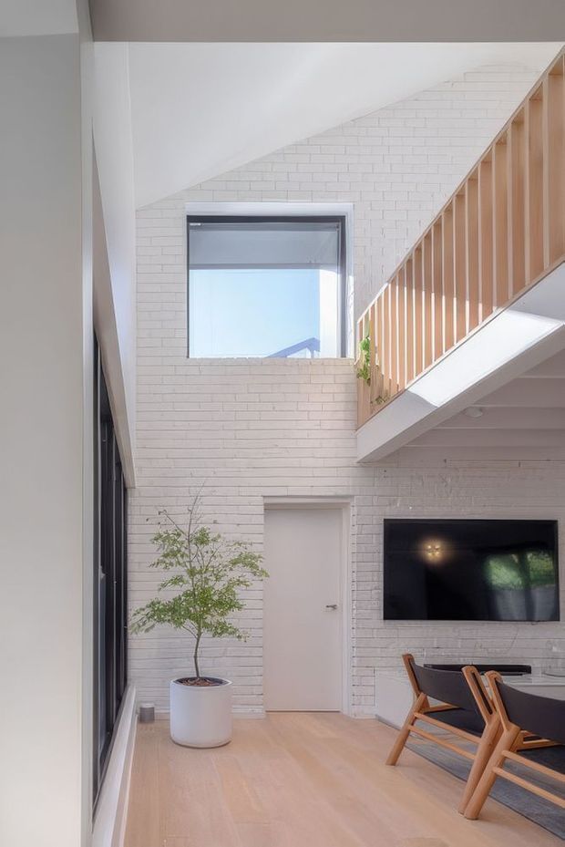 Bright contemporary double-height interior with white painted brick walls, pale timber flooring, and a timber-railed staircase leading to an upper level. A large window, potted indoor tree, wall-mounted television, and dining table with timber chairs are visible.