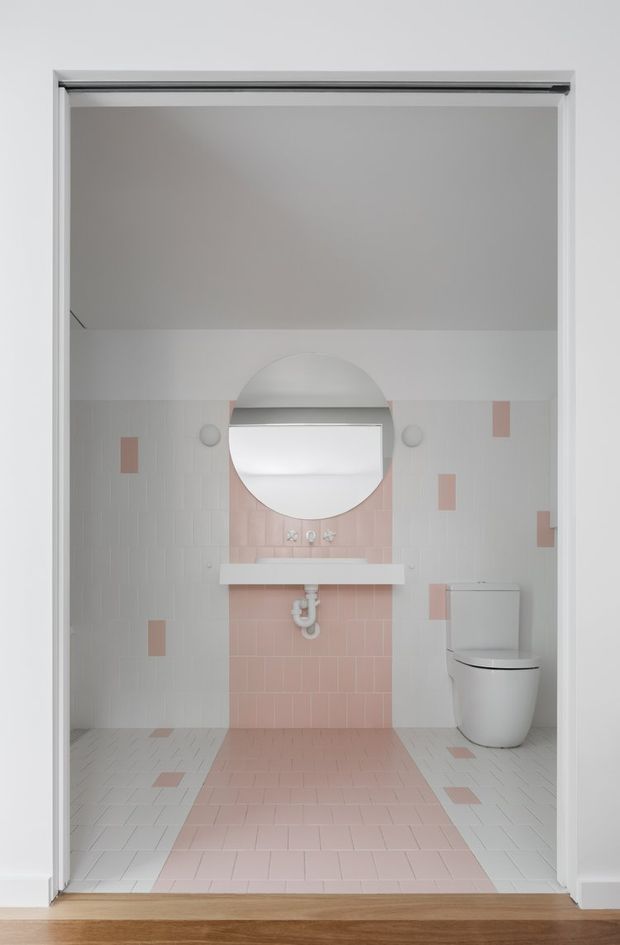 A bright, minimalist bathroom viewed through a wide doorway. The room features white and blush-pink tiled walls and floor, a wall-mounted white basin with exposed plumbing, a round mirror, and a modern white toilet. Small pink tile accents punctuate the predominantly white tile scheme, while a wood floor is visible outside the bathroom.