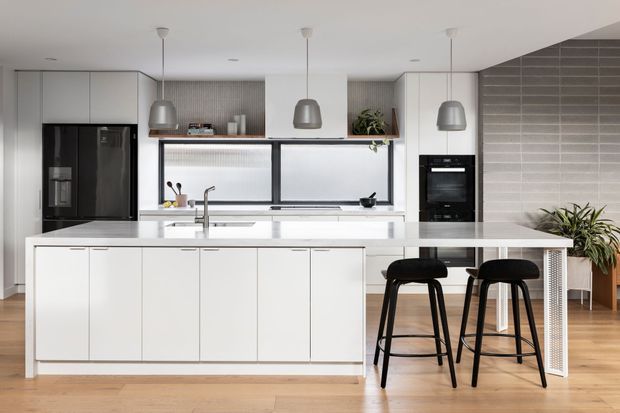 Bright contemporary kitchen featuring a large white island with integrated breakfast bar, handleless white cabinetry, dark appliances, a broad horizontal window, pendant lights, and a grey tiled feature wall. Warm timber flooring and indoor plants add contrast.