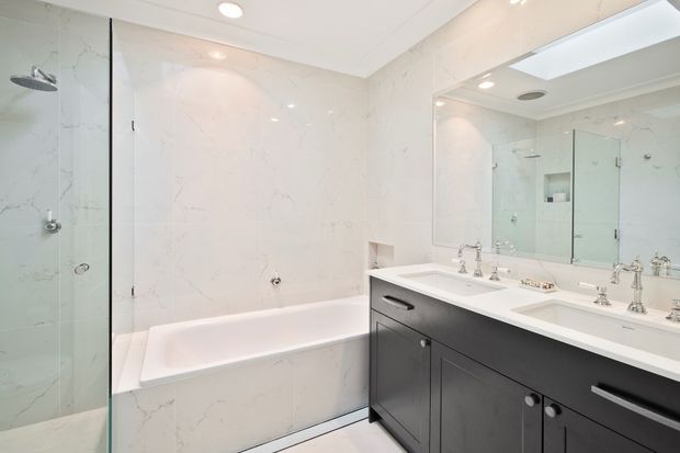 Bright, spacious contemporary bathroom with a white marble-look tiled bath and shower area, clear glass shower screen, large wall-to-wall mirror, and a dark charcoal double vanity with polished chrome fixtures. Recessed ceiling lighting and a skylight-like mirrored reflection add to the clean, refined feel.