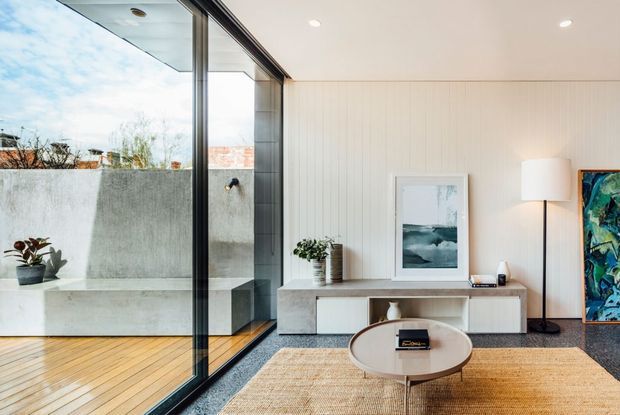 Bright contemporary living room with full-height black-framed glazing opening to a private terrace. The room features pale vertical wall panelling, a low light-toned built-in media console, neutral furnishings, a round coffee table, textured rug, floor lamp, and framed artwork. The terrace includes timber decking, a concrete privacy wall, and an outdoor shower fixture.