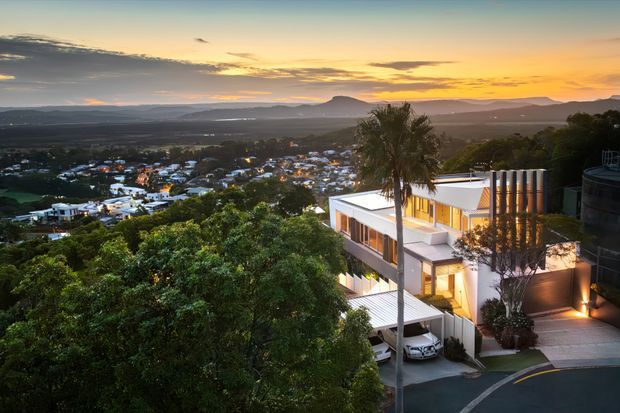 A contemporary multi-level residence on a steep, elevated suburban site, photographed at sunset. The home features crisp white rendered forms, extensive glazing, warm interior lighting, flat roof planes, balconies and terraces, a covered carport, and vertical architectural screening. Dense mature vegetation surrounds the property, with a broad suburban valley and hills visible beyond.