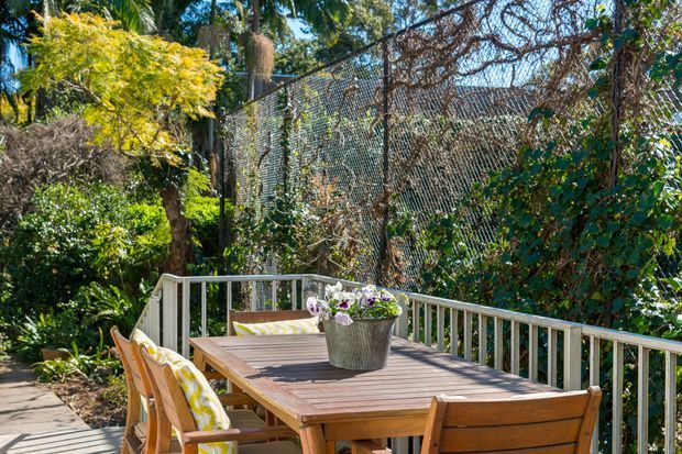A sunny outdoor dining deck surrounded by lush tropical garden planting and mature trees. A timber dining table with matching wooden chairs and yellow patterned cushions sits beside a white-painted railing, with a large mesh fence visible behind the vegetation.