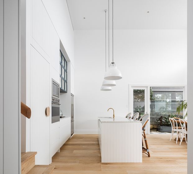 Bright, narrow open-plan kitchen and dining area with white floor-to-ceiling cabinetry, integrated appliances, a compact island with vertical panelling and a brass tap, timber flooring, pendant lights, and large glazed windows. A dining table and chairs are visible beyond the island. The space has a clean, refined contemporary coastal character with abundant natural light.