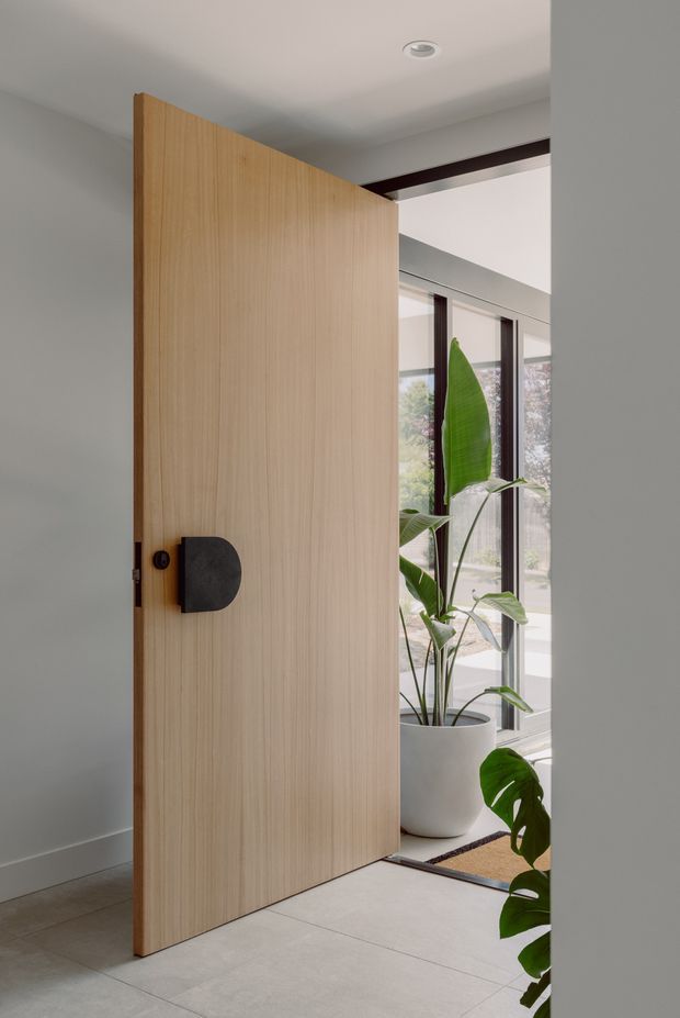 A contemporary entryway viewed from inside, featuring a tall pivot door with light vertical timber grain, a dark rounded handle, pale tiled flooring, white walls, and a bright glazed area beyond. Indoor tropical plants and dark structural or window-frame accents add contrast. The composition emphasises the oversized door and the connection between the entry and the light-filled adjoining space.