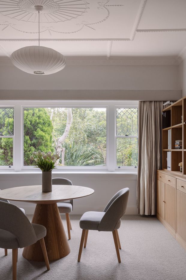 A refined dining room with a round sculptural timber dining table and upholstered dining chairs, positioned in front of large leadlight-style windows overlooking greenery. A built-in timber shelving and drawer unit occupies the right side, with full-length neutral curtains. The room features a decorative pressed ceiling with ornate moulded detailing and a suspended ribbed pendant light, creating a calm contemporary-traditional interior.