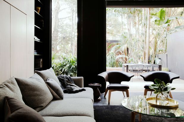 A refined contemporary living room with a large grey sectional sofa, sculptural dark lounge chairs, a glass coffee table, and black built-in shelving and cabinetry. Full-height black-framed glazing opens the room visually to a lush tropical garden and outdoor seating area. The space has a calm, sophisticated atmosphere with strong indoor-outdoor connection.