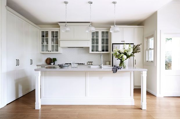 Bright, spacious white kitchen with a large island featuring paneled sides and decorative end supports, white shaker-style cabinetry, glass-front upper cabinets, integrated range hood, pale benchtops, pendant lights, and warm timber flooring. Natural light enters through the adjacent glazed door and windows.
