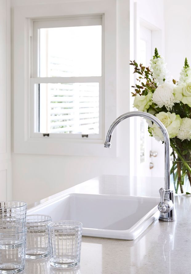 Bright, white kitchen sink area featuring a deep inset ceramic sink, polished chrome gooseneck faucet, pale stone countertop, white-framed window with plantation shutters, and a floral arrangement. Clear glass tumblers sit on the counter in the foreground.