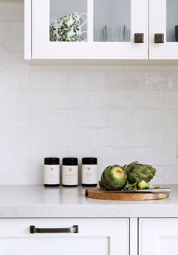 Close-up view of a bright white kitchen featuring glossy white subway-tile backsplash, white framed upper and lower cabinetry, dark metal cabinet hardware, and a pale benchtop. Glass-front cupboards display crockery and glassware, while a wooden cutting board with artichokes and a kitchen knife sits on the counter.