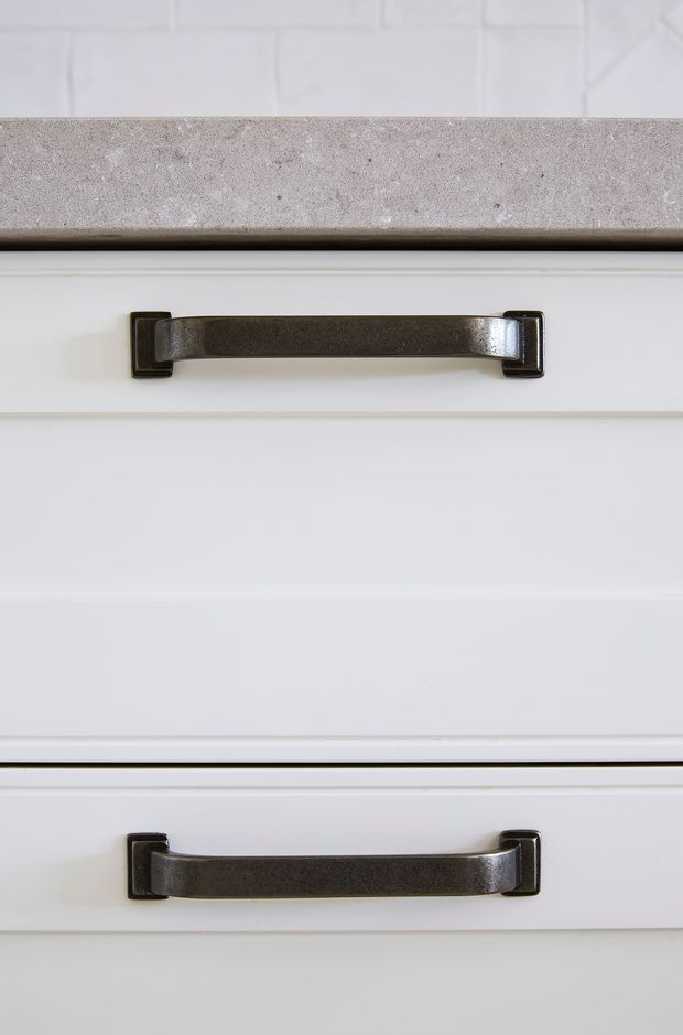 Close-up detail of white painted cabinetry with two dark aged-metal pull handles and a light grey stone benchtop above. The drawer fronts feature simple recessed panel detailing, creating a clean traditional-style kitchen or joinery finish.