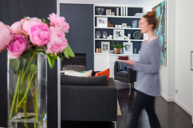 A stylish contemporary living room viewed from an adjacent area, featuring dark charcoal walls and flooring, a large dark sectional sofa with bright cushions, a built-in white display shelving unit filled with books and decor, and a linear fireplace set into the wall. A tall glass vase of pink flowers and a blurred person occupy the foreground.