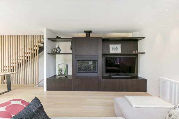 Bright contemporary living room featuring a full-height dark timber entertainment and fireplace joinery unit with open shelving, a television, and built-in fireplace. Pale timber flooring, white walls, recessed lighting, a light sectional sofa, and a vertical timber screen separating the stair or entry area create a clean, modern interior.