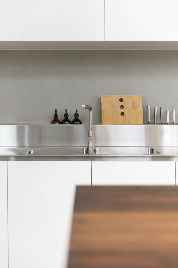 A close-up view of a minimalist kitchen with white handleless cabinetry, a stainless-steel double sink and tall mixer tap, a grey backsplash, and stainless-steel shelving or splashback detailing. Wooden chopping boards, dark bottles, and a knife block sit on the counter, while a warm timber island or benchtop occupies the foreground.