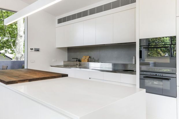 Bright, minimalist contemporary kitchen featuring a large white island with an integrated cooktop, a contrasting timber dining extension, white handleless cabinetry, a grey stone-look splashback, stainless-steel sink and appliances, and generous windows overlooking greenery.