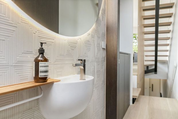 A compact contemporary bathroom with a wall-mounted white basin, brushed metal tapware, timber shelf, and textured white geometric wall tiles. A round mirror with concealed warm lighting sits above the basin. Through the opening, a timber stair with black vertical supports and pale built-in cabinetry is visible.